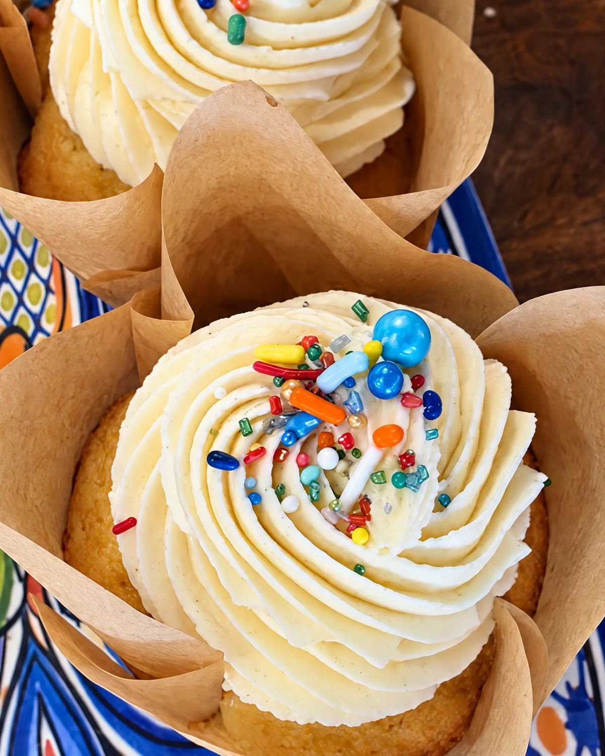 Vanilla cupcakes in paper wrappers, topped with swirled frosting and rainbow sprinkles