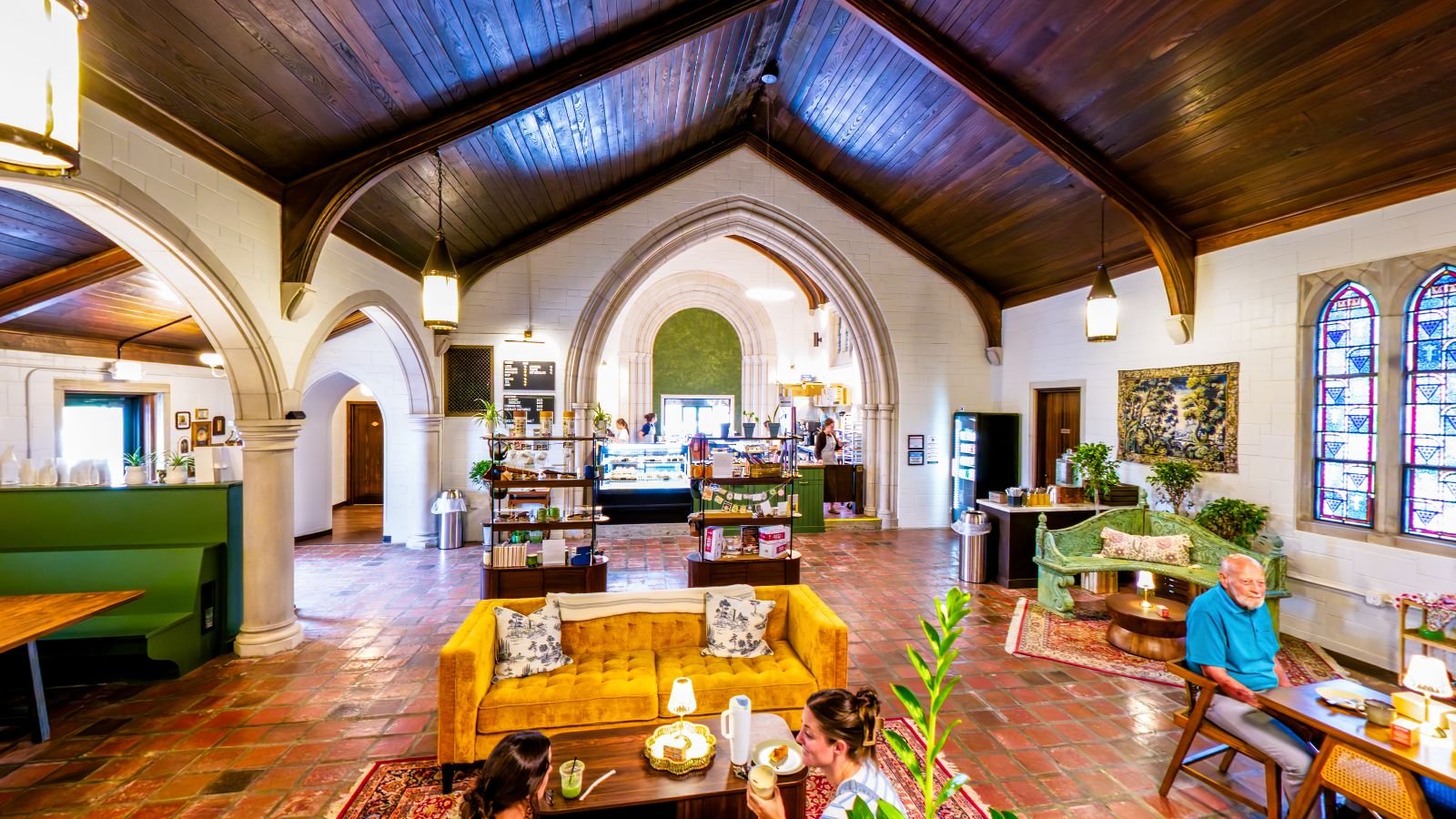 Inside Kairos Bakehouse: the pastry case under stone arches, a yellow velvet sofa, and a vaulted wood ceiling with hanging lanterns